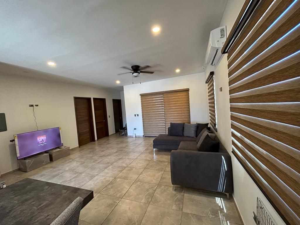 A spacious living area is shown, featuring a large sectional sofa and a television mounted on the wall. Light-colored tiled flooring complements the room, which has wooden blinds covering the windows. Air conditioning is visible, ensuring a comfortable environment.