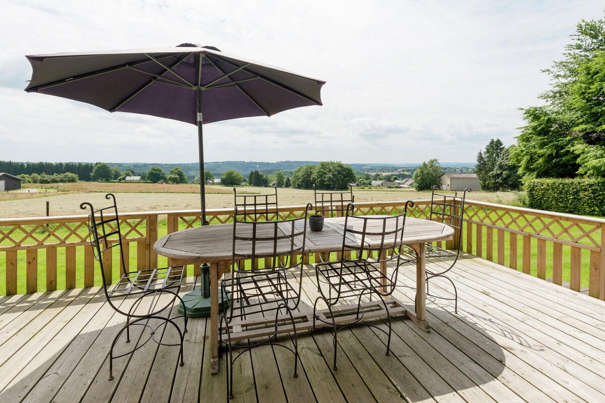A spacious outdoor terrace features a large wooden table surrounded by six metal chairs. An umbrella provides shade over the table, which overlooks expansive fields and distant hills. The wooden decking adds a natural touch to the serene landscape.