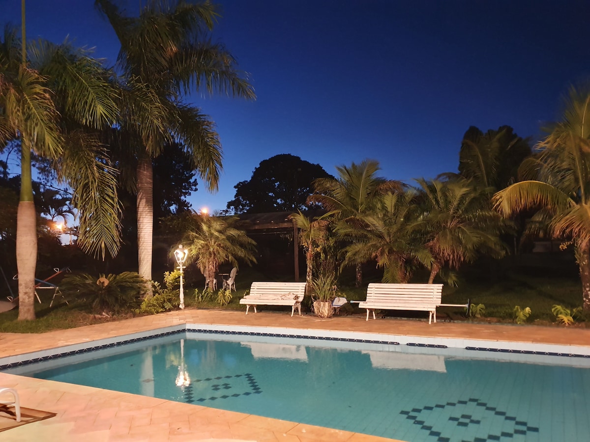 The pool area is surrounded by lush palm trees, with two white benches positioned nearby. The clear water of the pool reflects the evening sky, creating a serene atmosphere enhanced by soft lighting.
