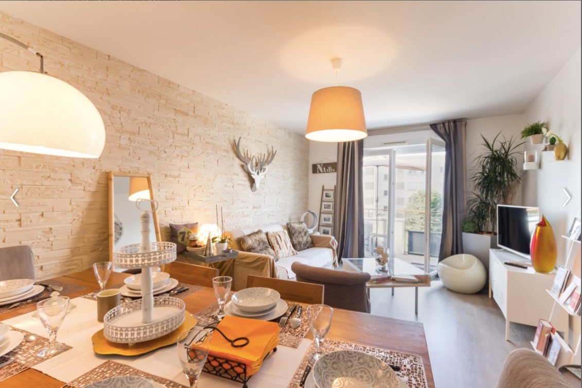 The living area is characterized by a light-colored wall adorned with decorative antlers. A comfortable seating arrangement is complemented by natural light flooding in through the glass doors that lead to the terrace. A dining table is set with elegant dishware, creating a welcoming dining space.