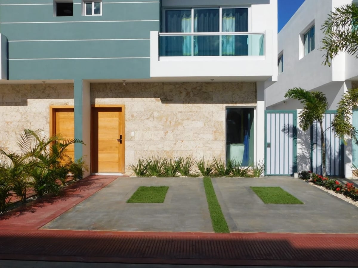 The exterior showcases a modern building with a light-colored stone facade and large windows. Two wooden doors provide access, while neatly arranged greenery and a small lawn contribute to the inviting entrance. A blue gate adds a touch of color and security.