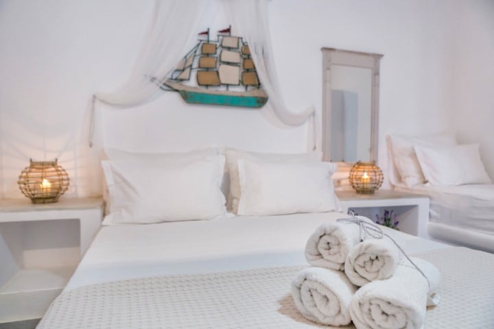 A spacious sleeping area features a neatly made bed with two pillows and decorative bed linens. Soft lighting is provided by two woven lanterns on either side. A handmade ship décor piece hangs above the bed, while neatly rolled towels are placed at the foot of the bed.