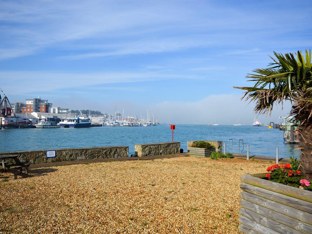 A waterfront view is presented, featuring a gravel area with seating and tropical plants. The water reflects the blue sky, and boats are visible in the distance, creating a serene coastal ambiance. The setting invites relaxation and enjoyment of the natural surroundings.