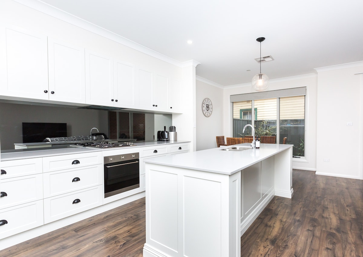 A modern kitchen is equipped with white cabinetry and a central island featuring a stone countertop. Sleek stainless steel appliances, including a stovetop and oven, are visible. Large windows allow natural light to illuminate the space, enhancing the inviting atmosphere.
