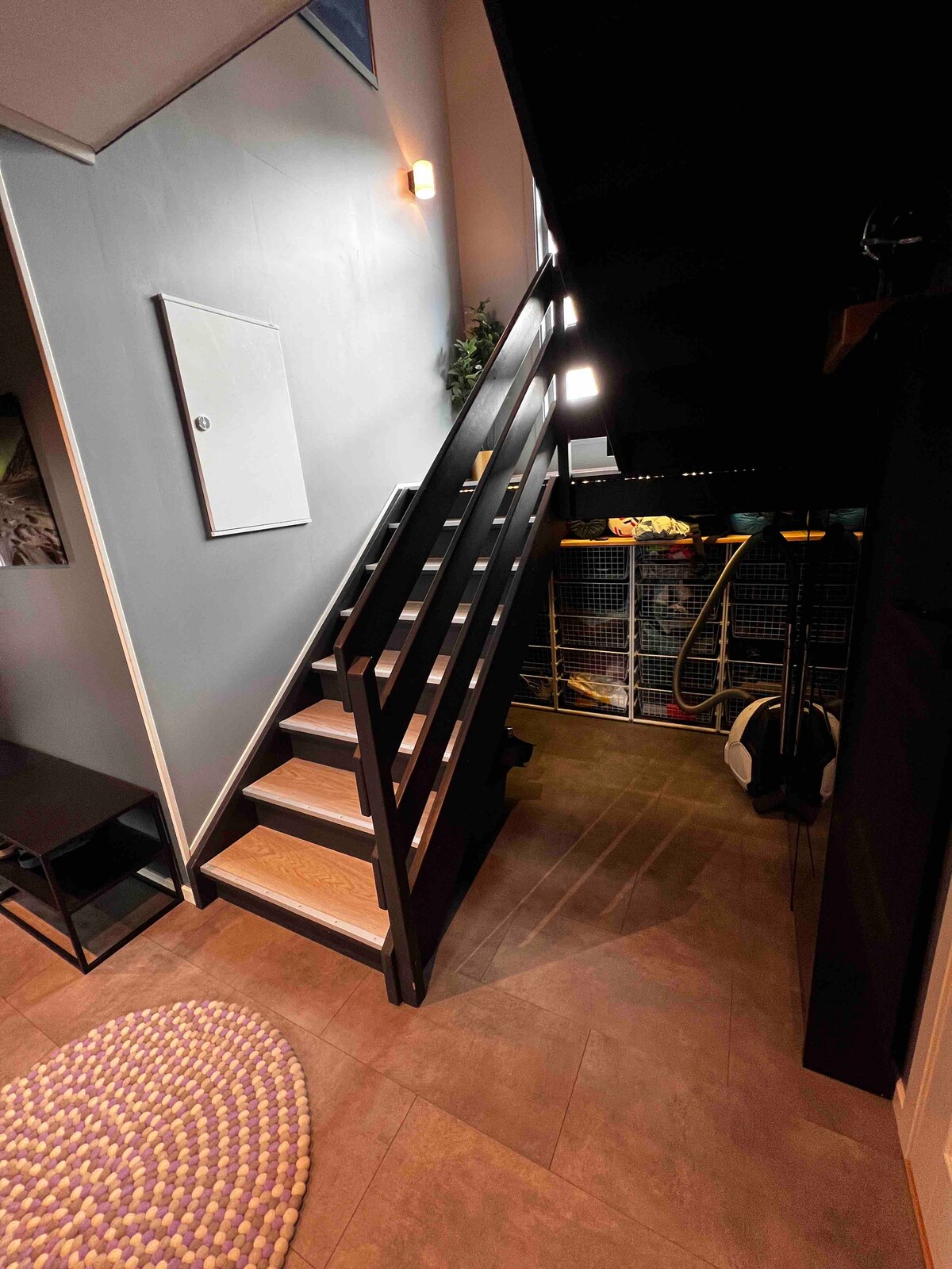 The entryway features a staircase with sleek black railings, leading to the upper levels. A small table is positioned to the left, while storage racks are visible in the background. Soft lighting highlights the modern design of the space.