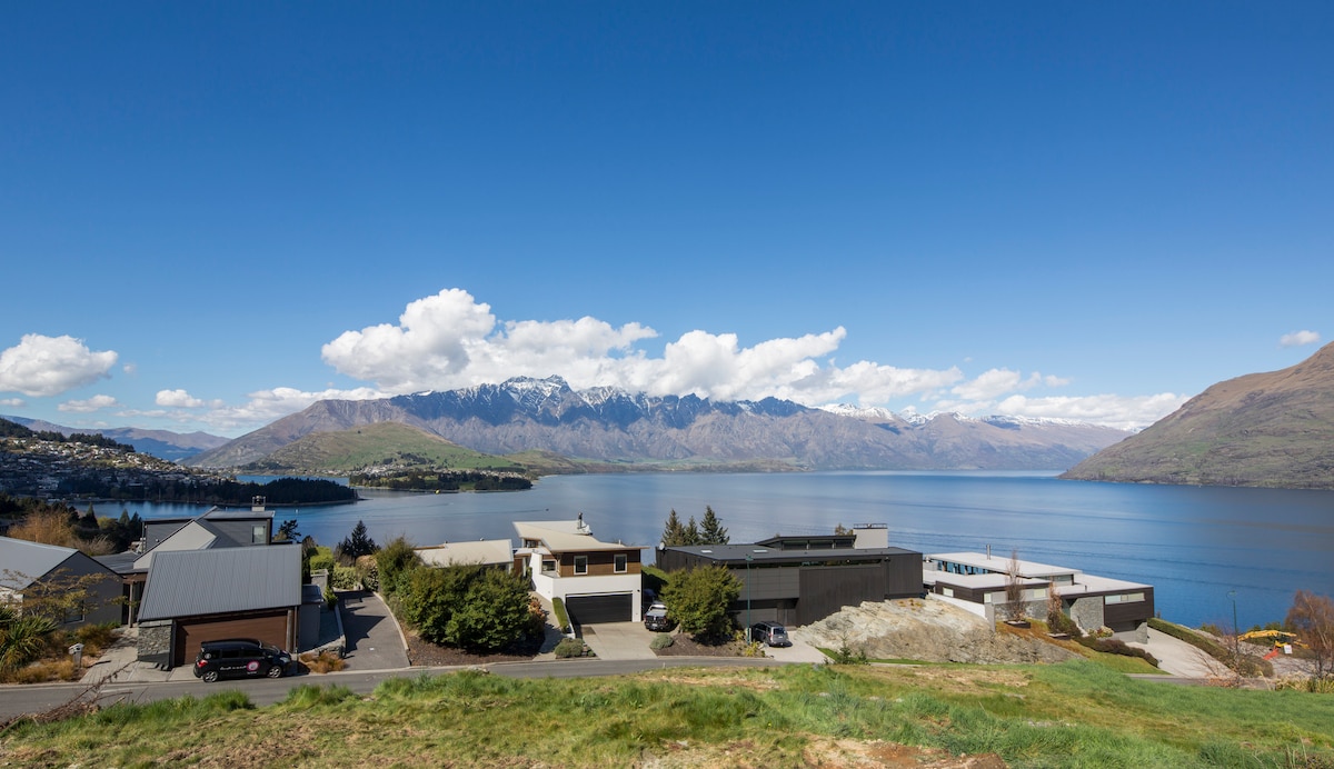 A panoramic view of Lake Wakatipu and The Remarkable, framed by distant mountains and a clear blue sky. The foreground features several modern homes with varying architectural styles, set against lush greenery and the calm water of the lake.