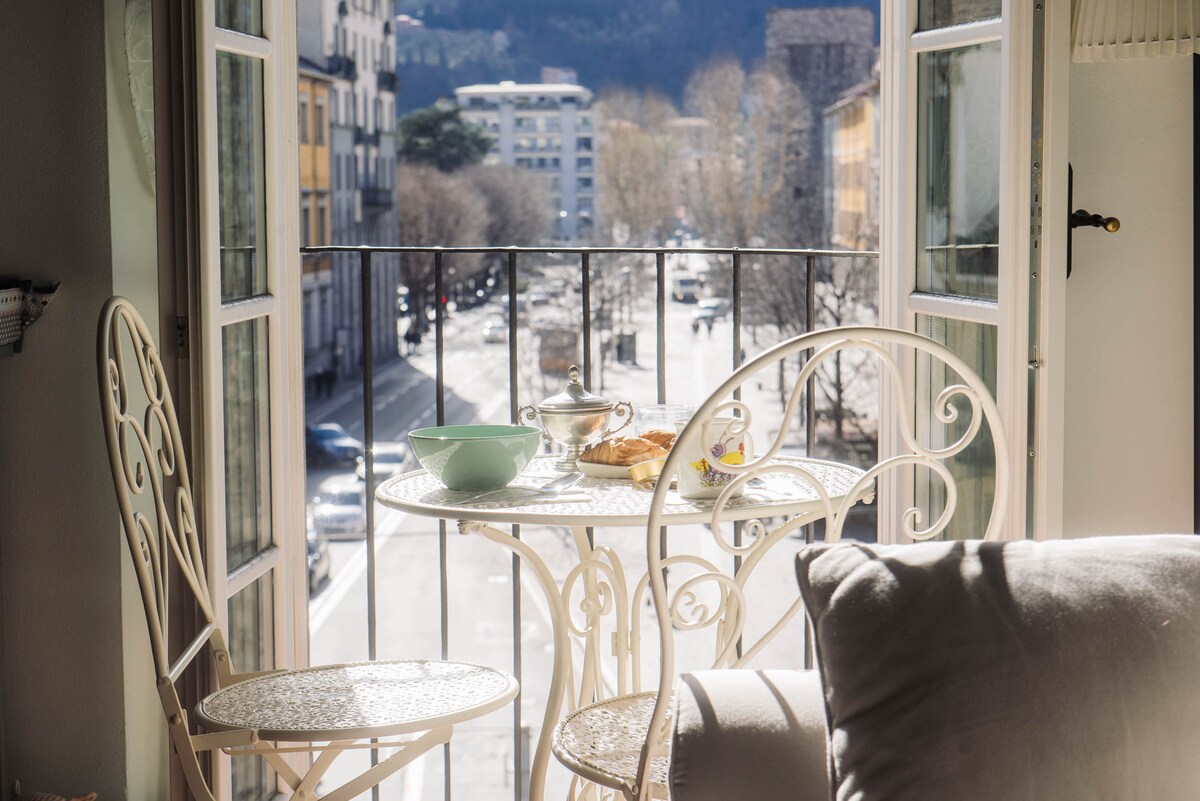 A small balcony overlooks a lively street, featuring a round table with two elegant chairs. On the table, a bowl and a teapot accompany a freshly baked pastry. Bright sunlight filters through open windows, creating a serene and inviting space.