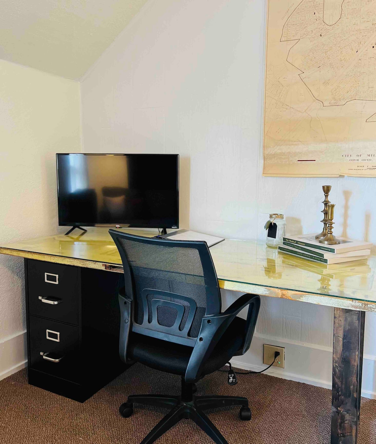 A designated workspace is highlighted by a sleek desk with a glass top and metal legs. A modern black office chair is positioned in front of a small computer screen. A filing cabinet with drawers, a decorative candle holder, and books are arranged neatly on the desk.