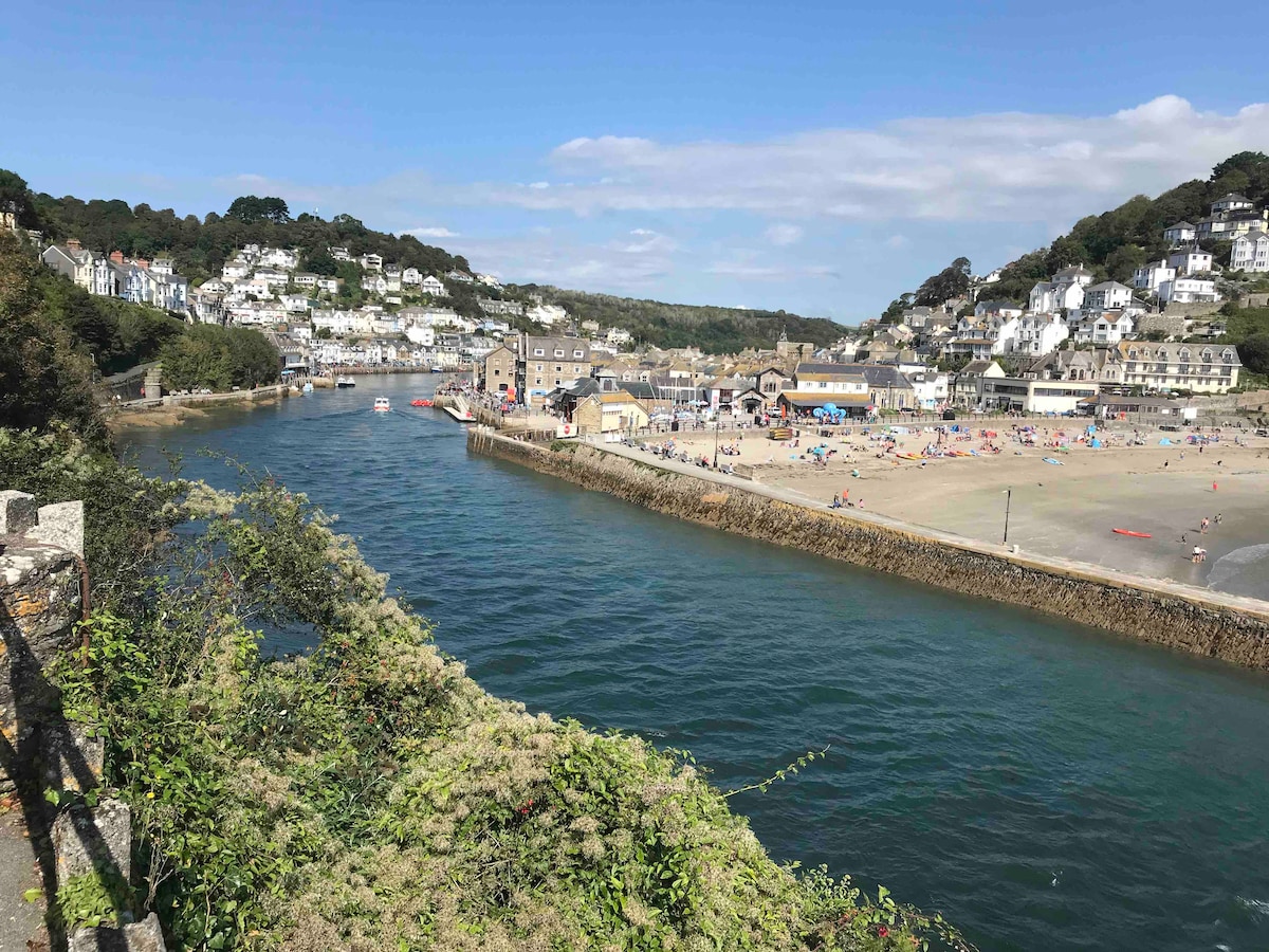 The image captures a scenic view of a tranquil harbor lined with sandy beaches, where numerous visitors can be seen enjoying the sun. The surrounding hills feature charming houses and green vegetation, creating a peaceful coastal atmosphere under a clear blue sky.