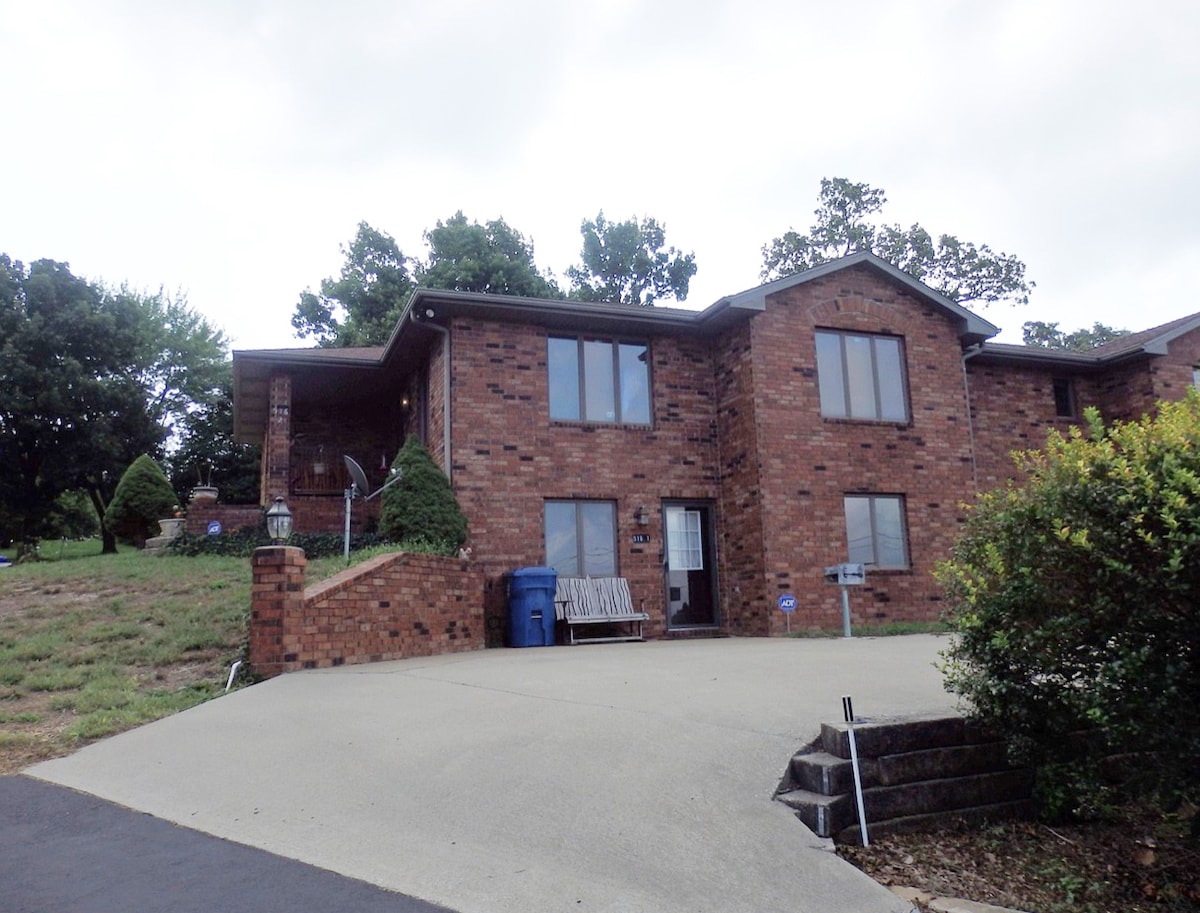 The exterior of a two-story brick house is visible, featuring large windows that reflect the surrounding greenery. A wide driveway leads to the entrance, where a wooden bench sits. The yard includes manicured bushes and trees, providing a quiet, inviting atmosphere.