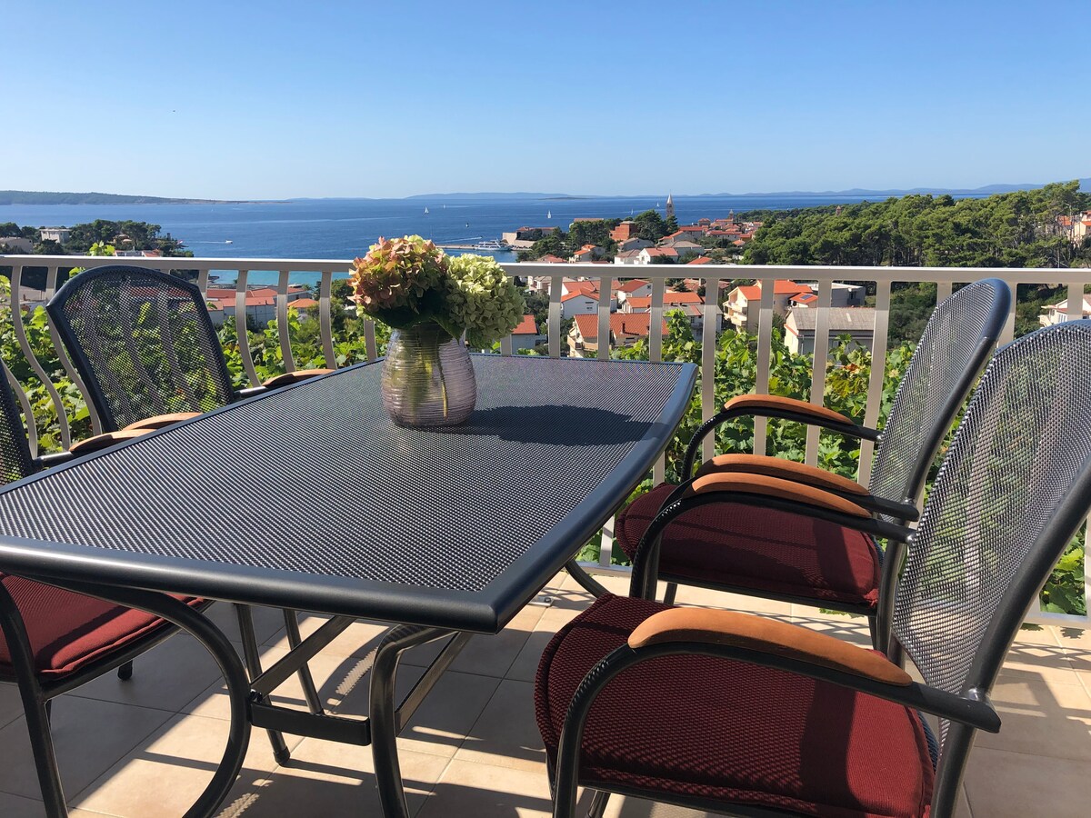 A terrace table is surrounded by four chairs, offering a scenic view of the sea and nearby town. A vase with flowers is positioned at the center of the table, while the clear blue sky adds to the tranquil atmosphere.