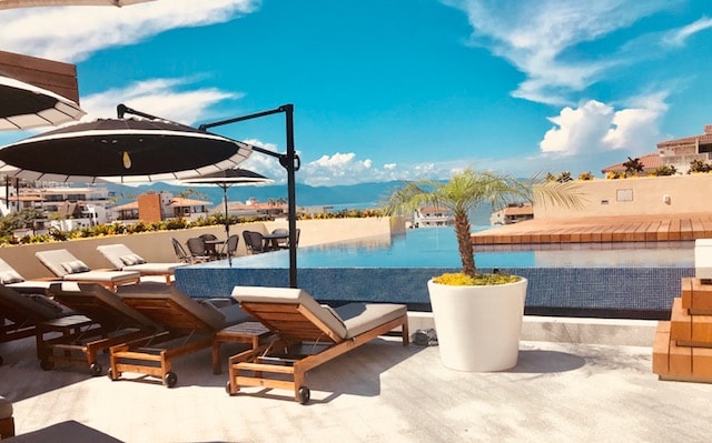 A rooftop pool area is presented, featuring lounge chairs arranged around the infinity pool. A potted palm adds a touch of greenery, while umbrellas offer shade. The backdrop showcases a clear blue sky and distant mountains, creating a serene outdoor space.