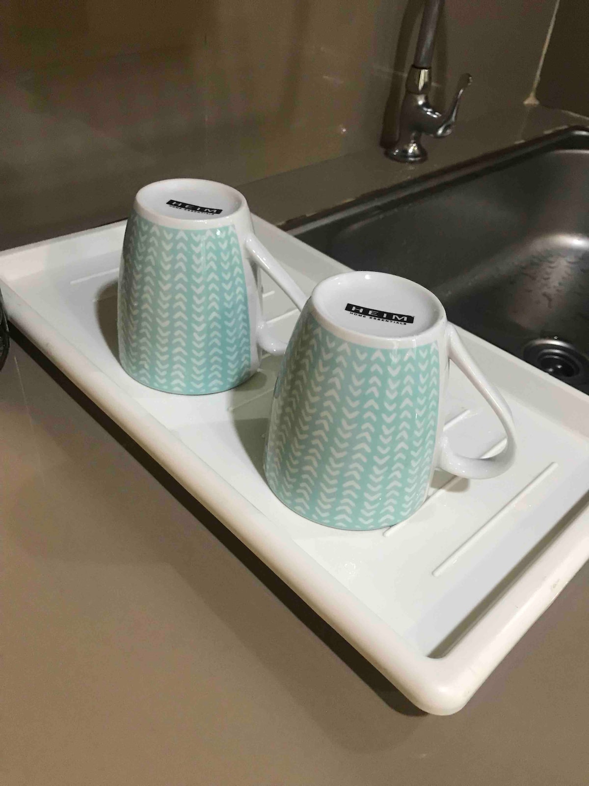 Two ceramic mugs with a subtle turquoise geometric pattern are placed on a white drying tray. The tray sits next to a stainless steel sink, reflecting a clean and tidy kitchen environment.
