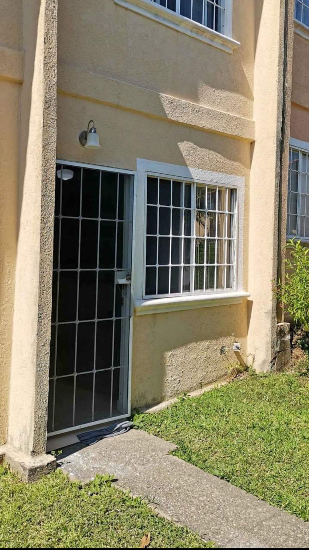 A welcoming entrance is visible, featuring a metal security door framed by a small path of concrete leading to a grassy area. The adjacent window, adorned with multiple panes, allows natural light to brighten the entrance, displaying a simple and functional design.