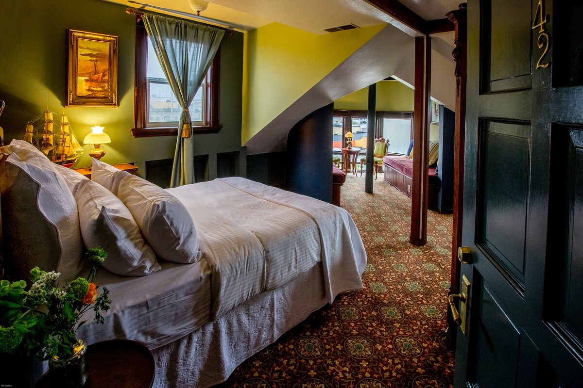 A cozy room features a spacious bed with crisp white linens. The rich green walls are paired with warm lighting from a bedside lamp. A seating area with comfortable furniture is visible in the background, while a window provides natural light and views of the surroundings.