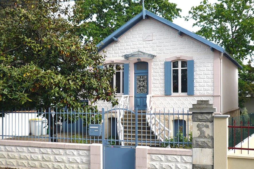 The exterior of a charming two-story house is displayed, featuring a light stone facade with blue trim. White railings surround the entrance steps. Large windows with blue shutters allow natural light to enter. A well-maintained garden and a gated fence enhance the inviting appearance.