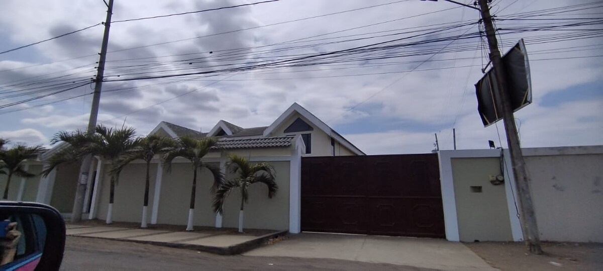 A modern residence is enclosed by a low wall, featuring a large, dark gate. Palm trees are planted along the front, and a multi-gabled roof is visible above the gate, with soft clouds partially covering the blue sky.