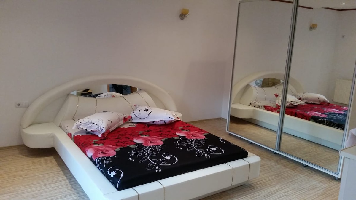 A modern sleeping area features a unique curved bed frame with a floral design on the bedding. A spacious mirrored wardrobe reflects the room's soft lighting, while light-colored wooden flooring complements the neutral wall tones.