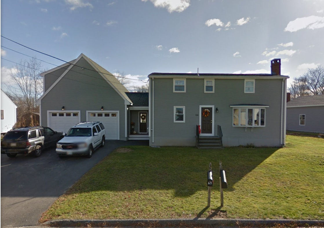 A two-story house features a gray exterior with large windows and a front porch. Two vehicles are parked in the driveway, with additional parking space available. The front yard is neatly maintained, showcasing a grassy area and steps leading to the entrance.