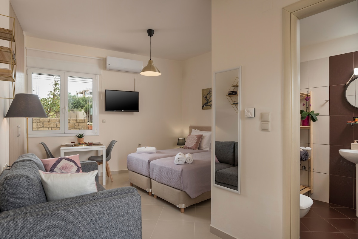 A modern studio layout is featured, showcasing a cozy sleeping area with a spacious bed covered with neatly arranged towels. A dining table is positioned near a bright window, and a comfortable sofa provides additional seating. A wall-mounted TV and a mirror enhance the room's functionality.