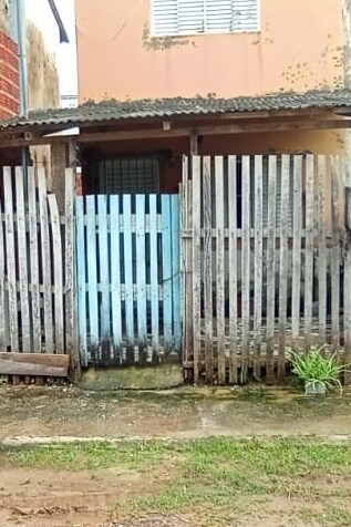 A wooden picket fence surrounds the entrance to a modest property, featuring a blue gate. The weathered facade of the house is visible behind the fence, with a small window partially obscured by a screen. A patch of grass and damp ground are seen in the foreground.
