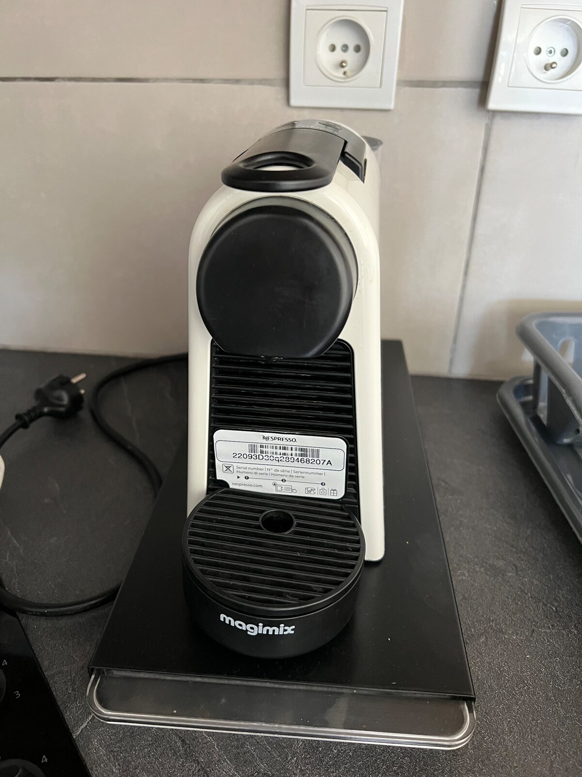 A Nespresso coffee machine is positioned on a countertop, featuring a sleek white design. The machine is connected to a power source visible at the back, with a black drip tray underneath. A small label is affixed to the front, providing details about the appliance.