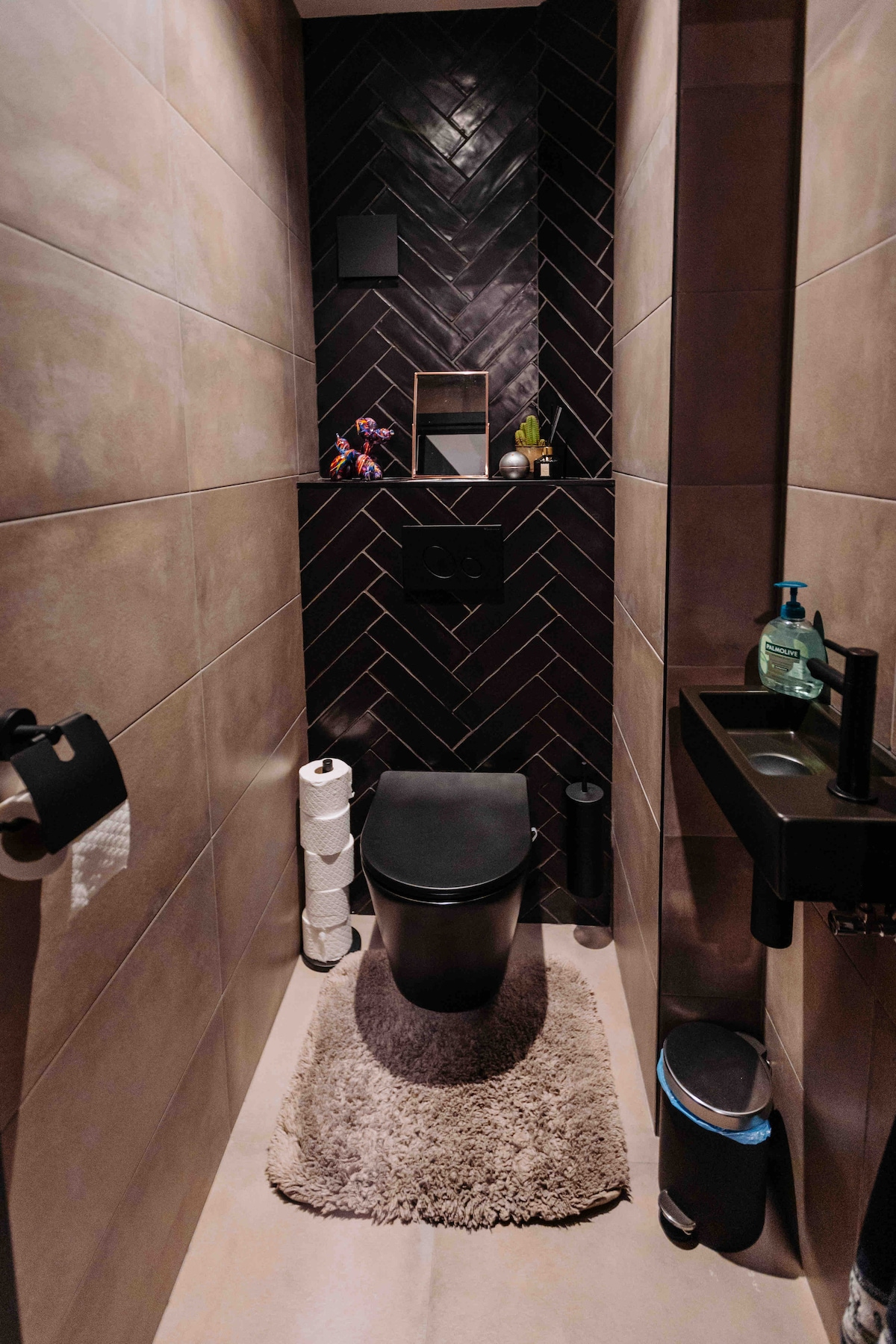 The bathroom features a modern toilet positioned against textured wall tiles. A small shelf holds toiletries, and a round mirror is mounted above. A plush, light-colored rug lies on the floor, adding comfort to the space.