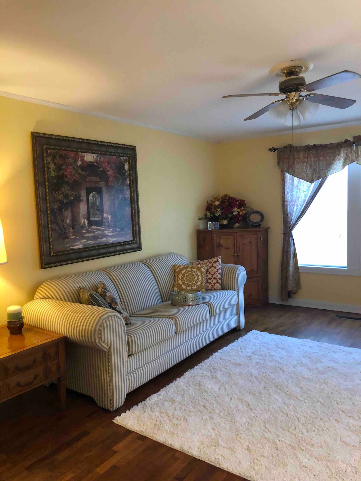 A cozy living space features a striped sofa adorned with decorative cushions, positioned next to a wooden cabinet. A small lamp provides soft lighting, while a colorful wall painting enhances the ambiance. Natural light filters through a nearby window, covered by sheer curtains.