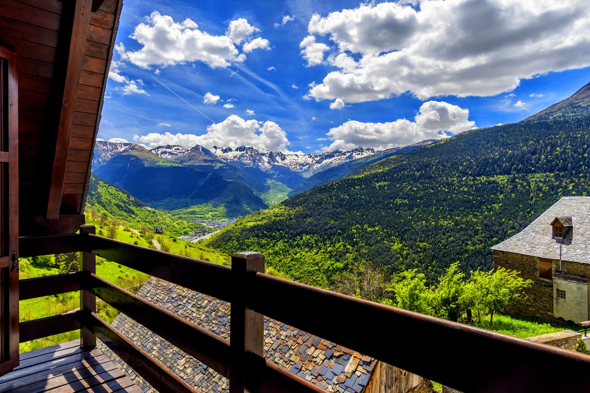 A wooden balcony offers expansive views of the surrounding mountains and valleys. Snow-capped peaks are visible in the distance, while lush greenery adorns the foreground under a bright blue sky with fluffy clouds.