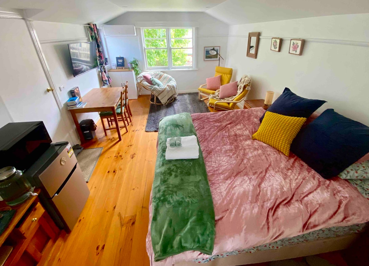 The loft room features a spacious bed adorned with colorful pillows and a soft throw. A small dining table is positioned nearby, complemented by a cozy armchair. Natural light fills the room through the windows, which overlook greenery, creating a bright and airy atmosphere.