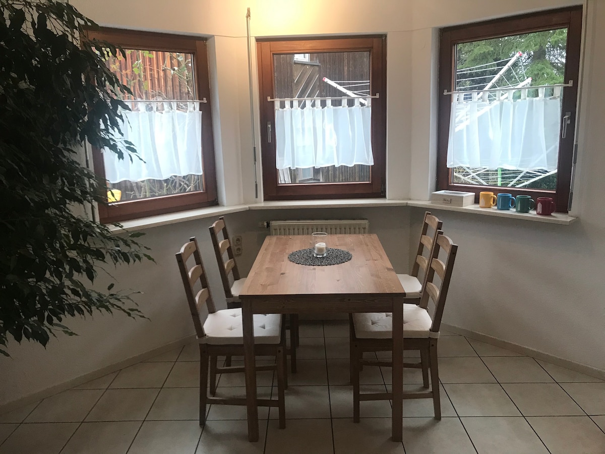 A light-filled dining area features a wooden table surrounded by four chairs. Three windows draped with sheer white curtains provide natural light, enhancing the room's openness. A small decorative centerpiece is positioned on the table, adding a subtle touch to the inviting space.