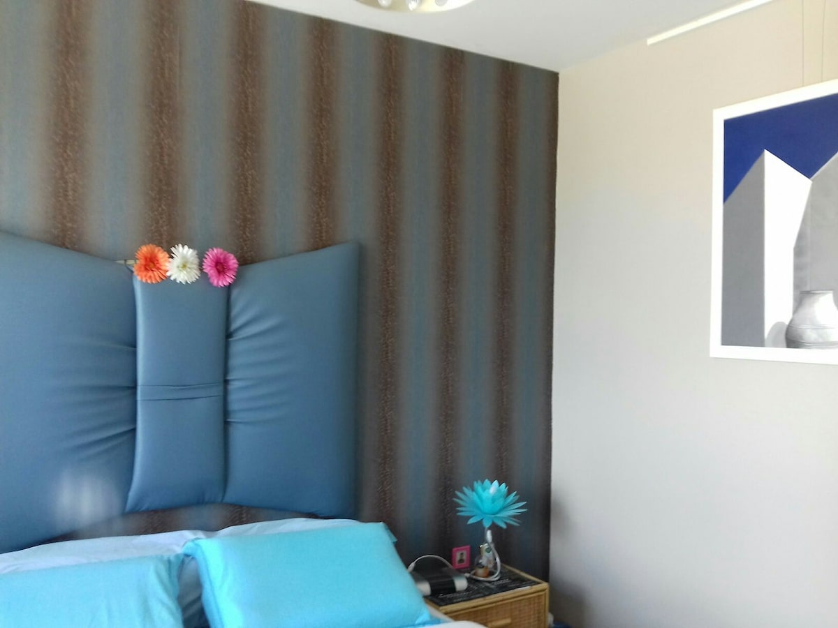 A comfortable sleeping area features a large padded headboard in a deep blue tone. Decorative flowers are placed across the top of the headboard. A bedside table holds a small blue decorative item, alongside crisp blue bedding, against a softly patterned wall.