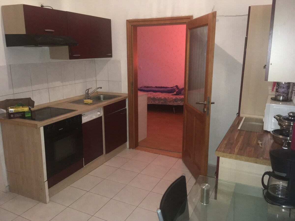 A compact kitchen features dark cabinetry and light countertops, equipped with a stove, oven, and dishwasher. A doorway leads to a softly lit bedroom in the background. The tiled floor extends throughout the space, contributing to the overall functionality.