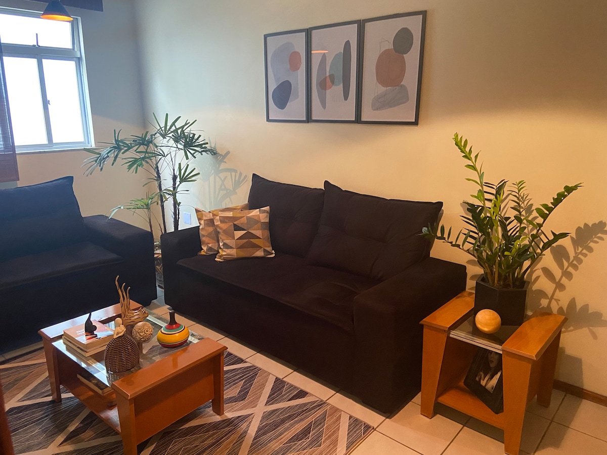 A cozy living area features two dark sofas arranged around a wooden coffee table. Decorative items, including a plant and various artworks, add character to the space. Natural light floods in through a nearby window, enhancing the warm atmosphere.
