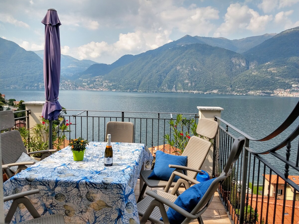 A terrace is featured with a dining table draped in a blue and white patterned cloth. Several comfortable chairs surround the table, with a bottle of champagne on display. The panoramic view of Lake Como and surrounding mountains is visible in the background, providing a scenic backdrop.