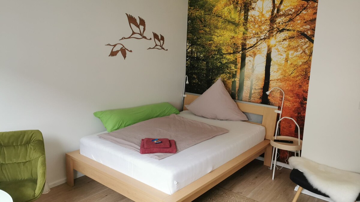 A stylish bedroom features a wooden bed with a white mattress set against a vibrant wall mural of a forest in autumn. A light green pillow and a mauve comforter are positioned on the bed. A cozy armchair and a small side table are also present in the room.