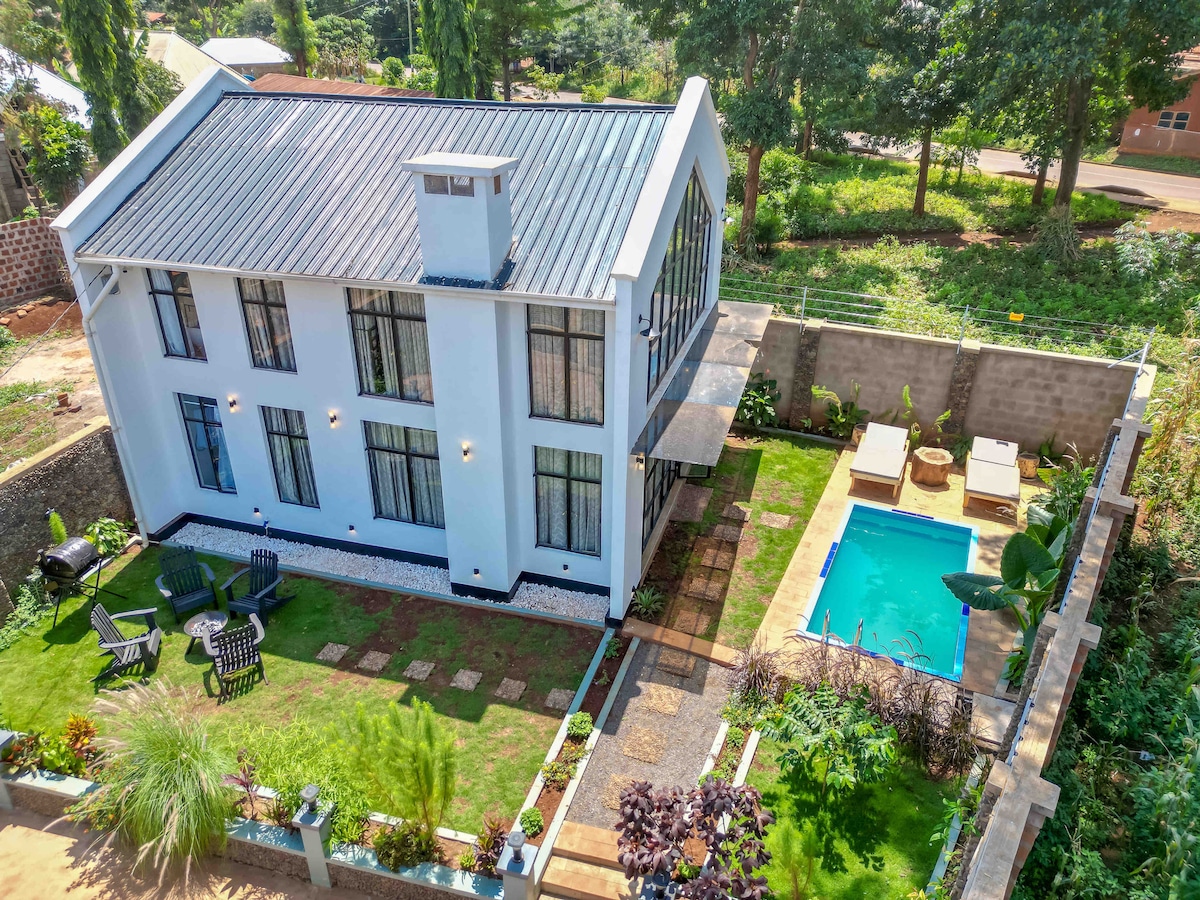 An aerial view showcases a modern two-story house with large windows, surrounded by a well-maintained garden. A private pool is positioned alongside the house, with lounge chairs situated nearby. Pathways of stone lead through the lush green lawn, enhancing the outdoor space.