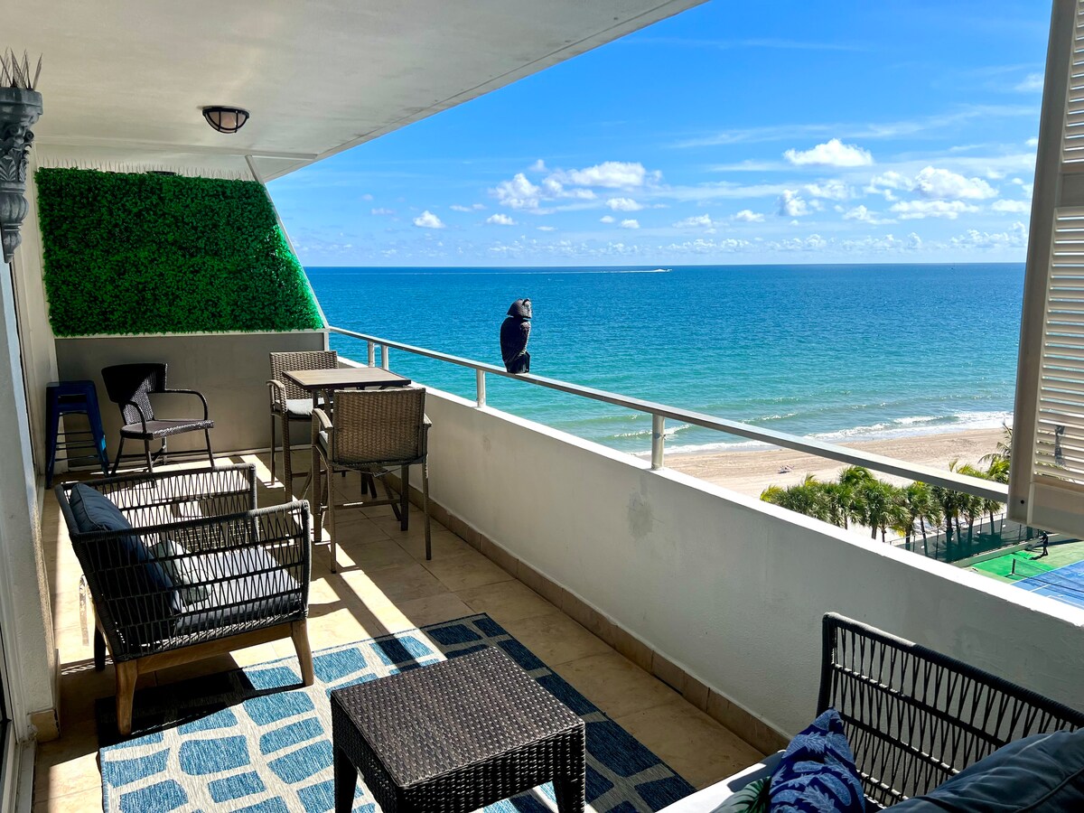 A large balcony offers ocean views with seating arrangements featuring woven chairs and a low table. The flooring is tiled, and a lush green wall adds a natural touch. The unobstructed view shows turquoise waters meeting a sandy beach under a clear blue sky.