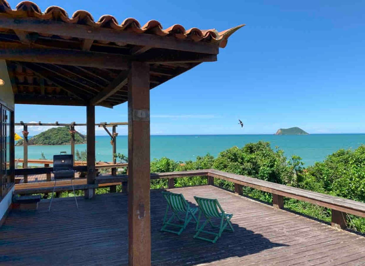 A spacious wooden deck overlooks a sweeping view of the ocean and nearby islands. The deck features two green lounge chairs and a barbecue area, framed by lush greenery. The clear blue sky and calm waters create a serene coastal atmosphere.