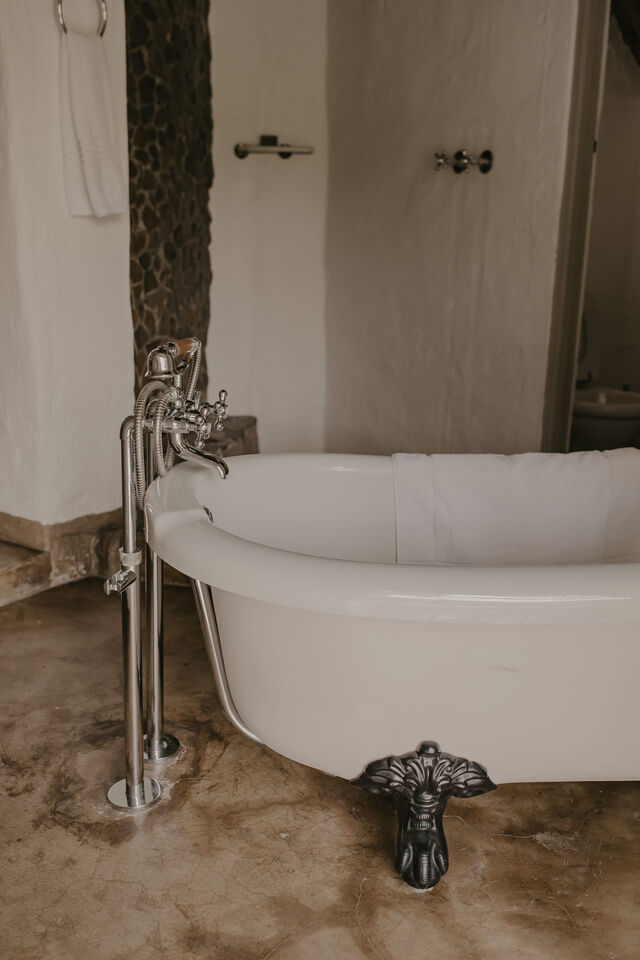 A freestanding bath with a distinctive clawfoot design is positioned next to a textured stone wall. It features a chrome faucet with dual handles, and a soft white towel drapes over the edge of the bath. The floor displays a polished concrete finish.