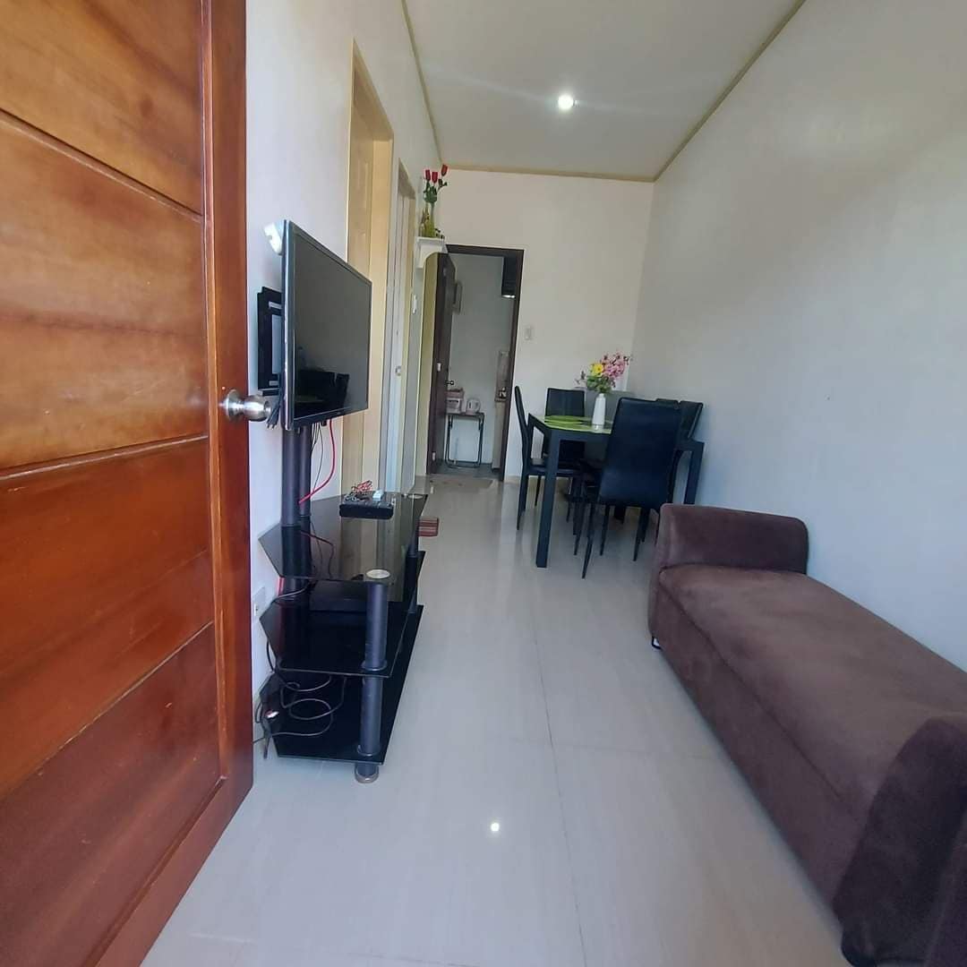 A narrow hallway leads to a living area featuring a brown sofa and a TV mounted on a stand. A dining table with black chairs is positioned nearby, complemented by a soft glow from overhead lighting. A door opens to an adjoining room.