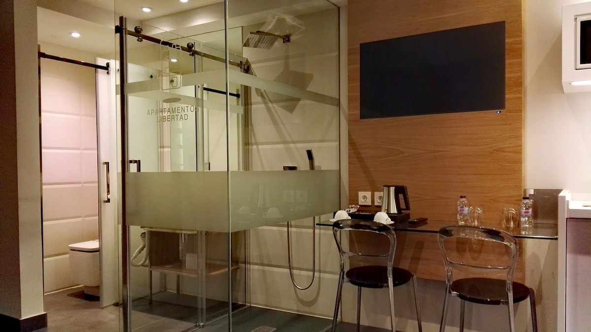 A modern suite featuring a glass-enclosed bathroom with a rainfall shower and a toilet. A small dining area with two transparent chairs is positioned next to a wooden desk. A flat-screen television is mounted on the wall, complementing the sleek design.