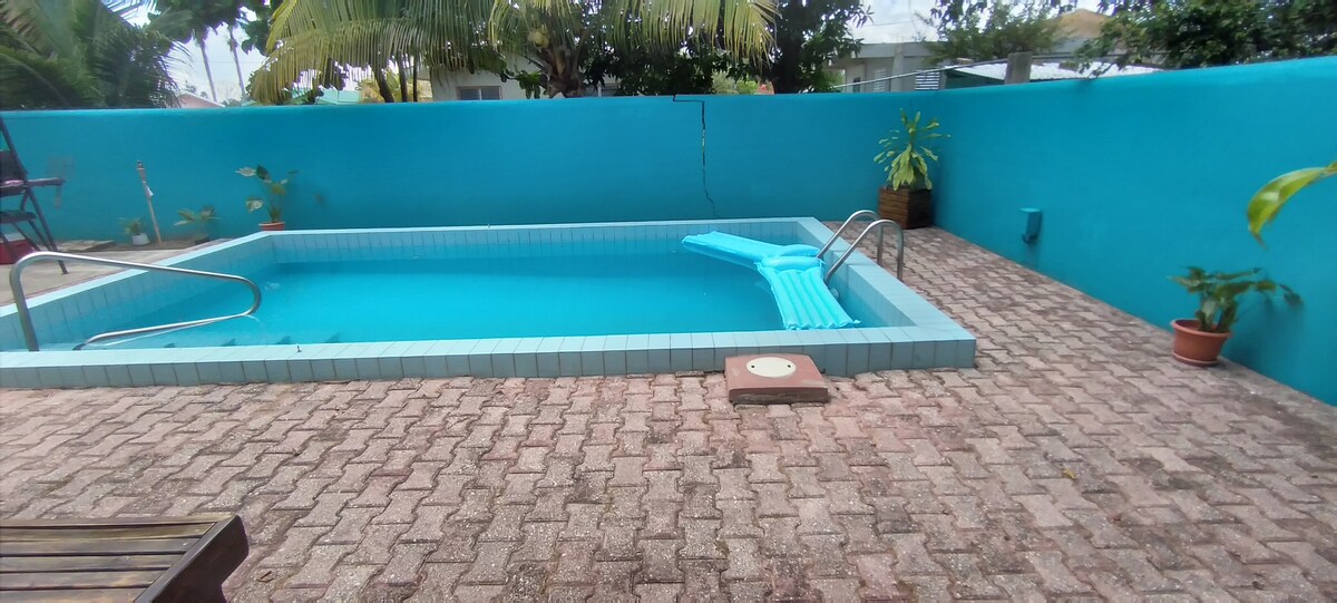 A private inground pool with a gradient depth of 4 to 6 feet is set within a turquoise-walled backyard. Surrounding the pool, natural stones pave the area, with a lounging section featuring beach chairs. Potted plants are positioned nearby, enhancing the outdoor setting.