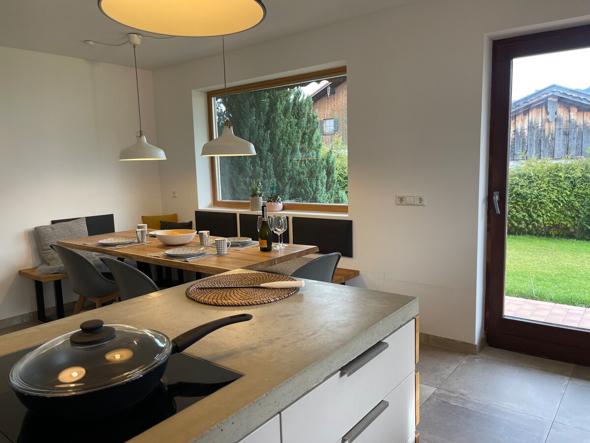 A modern kitchen area features a spacious island with a stovetop and sleek counter. A large window offers views of greenery, while a dining table with seating for six is illuminated by pendant lights. A door leads to the garden beyond.