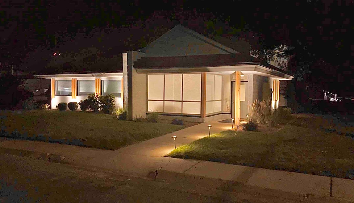 The exterior of the home is softly illuminated at night, showcasing a modern design with large windows and a welcoming front entry. A clear path leads to the entrance, surrounded by landscaped greenery and subtle outdoor lighting that enhances the architectural features.