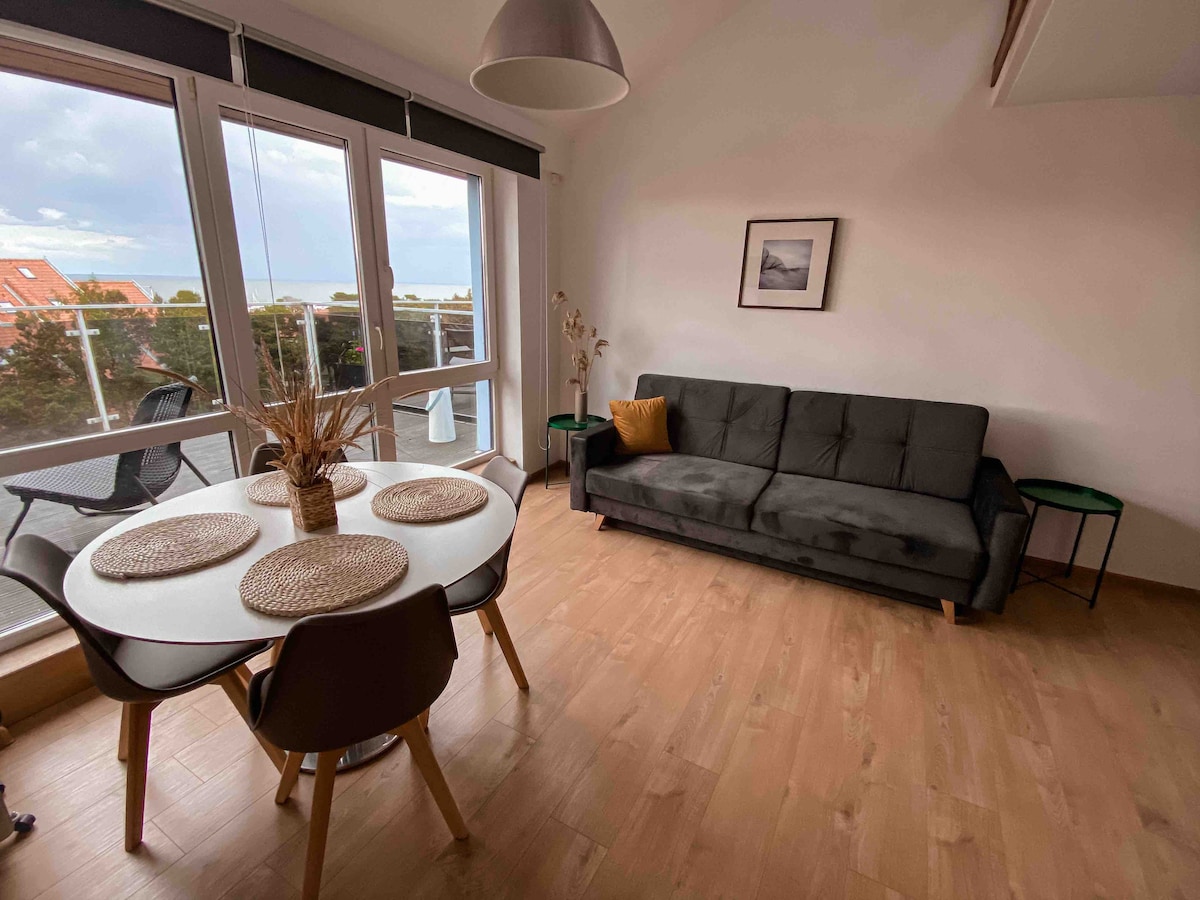 A cozy living area is furnished with a dark sofa and a round dining table set for four. Natural light filters through large windows, offering views of the outdoor space. A minimalist decorative piece rests on a side table, adding a touch of elegance.