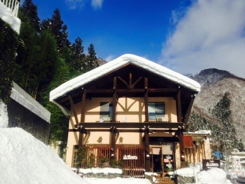 The building features a distinct Nordic architectural style, with a steeply pitched roof covered in snow. Large windows allow for natural light, offering views of the surrounding mountains. The entrance is framed by snow-laden shrubs, creating a serene winter atmosphere.