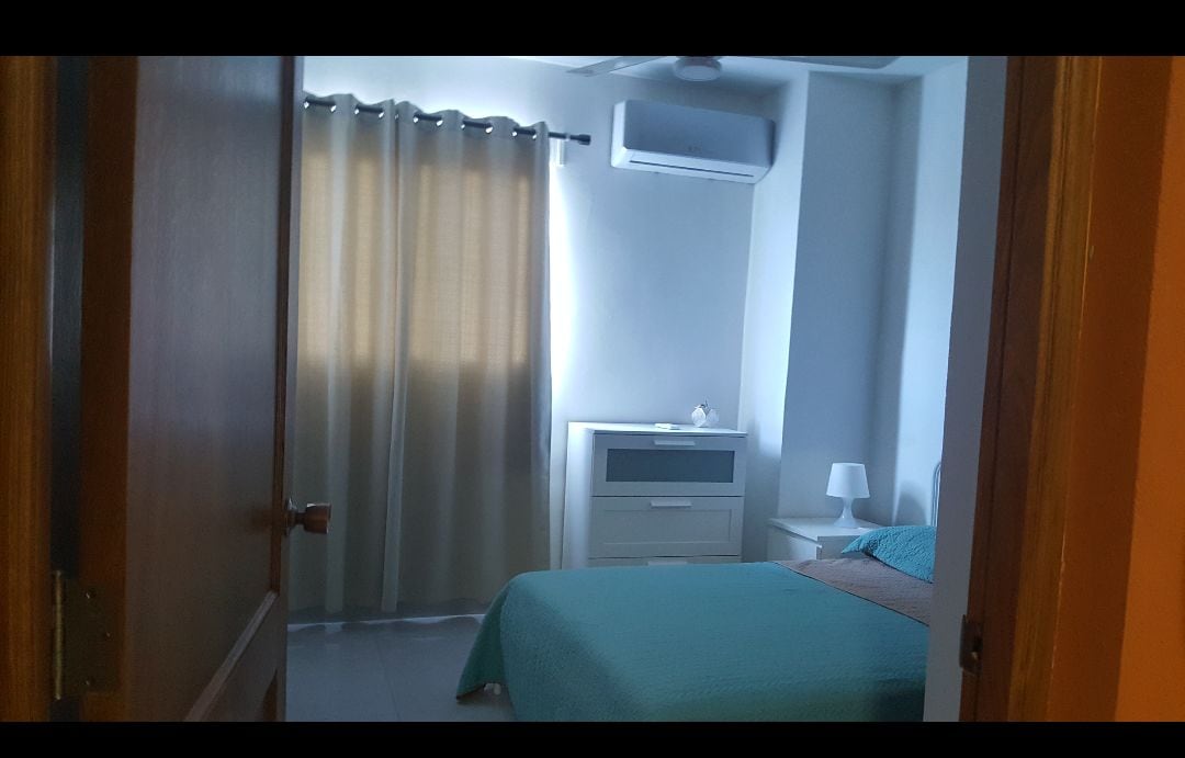 A serene bedroom is visible, featuring a spacious bed covered in a light turquoise blanket. A soft light illuminates the room from a nearby lamp on a nightstand. Curtains gently filter sunlight, and a modern dresser adds functionality.