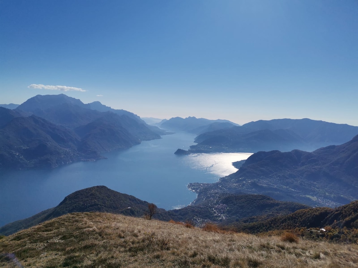 A scenic view of Lake Como is presented, with mountains framing the shoreline. The lake's surface reflects sunlight, creating a shimmering effect. The background features distant peaks, while the foreground shows grassy terrain. Clear skies enhance the overall atmosphere of tranquility.