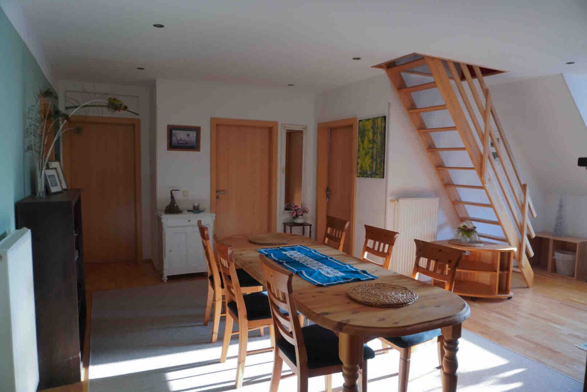 A well-lit dining area features a wooden table surrounded by six chairs. A staircase leads to the upper floor. The room includes two closed doors and a sideboard, enhancing the approachable layout. Natural light floods through the windows, creating a welcoming space.